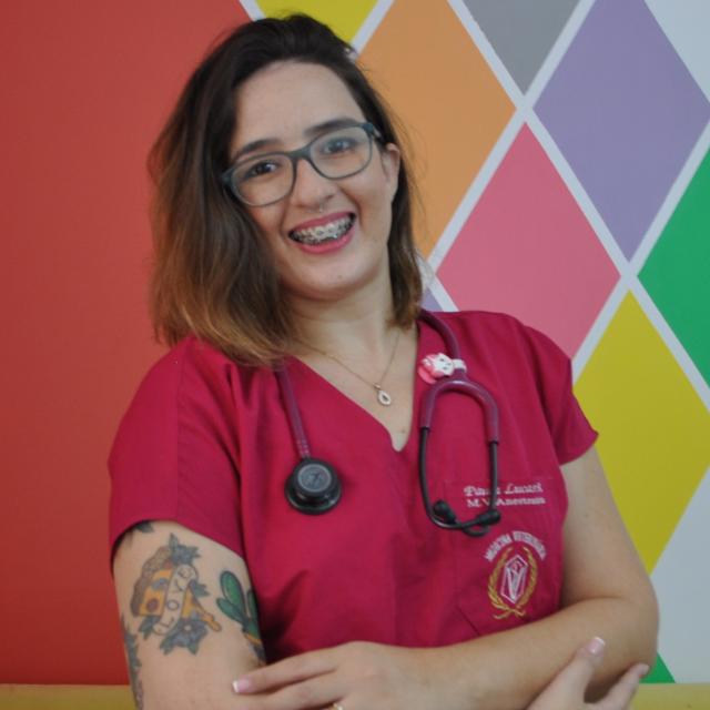 Foto da Médica Veterinária Paula, ela tem cabelos lisos de comprimento médio, está sorrindo e com os braços levemente cruzados. Veste um pijama cirurgico na cor bonina e usa um estetoscópio no pescoço